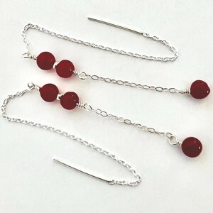 May include: A pair of silver threader earrings. Each earring features three red beads and a single red bead at the end. The earrings are shown against a white backdrop.