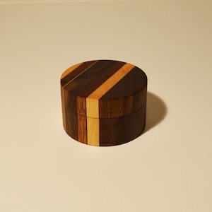 May include: A small, round wooden box with a striped pattern in shades of brown and light wood. The box is made of different types of wood, creating a unique and decorative design.
