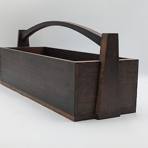 May include: A rectangular dark brown wooden caddy featuring a long, arched handle that extends upward from the center of the two shorter sides. The box has visible joinery at the corners and a smooth, stained finish with a deep wood grain texture.