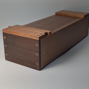 Peut inclure: Une boîte rectangulaire en bois, avec un corps brun foncé et des accents plus clairs sur le couvercle. La boîte est construite avec des vis apparentes et présente un design simple et élégant. Elle est probablement utilisée pour le rangement.