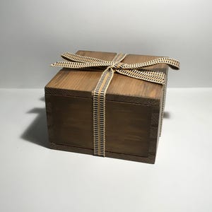 Puede incluir: Una caja de madera cuadrada con un acabado marrón oscuro, atada con una cinta decorativa. La cinta es beige con rayas verticales negras y doradas. La caja está cerrada y parece estar vacía.