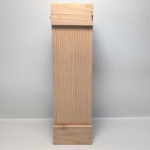 May include: A tall, rectangular wooden box with a natural finish. The box is constructed from light-colored wood, featuring visible grain patterns. The top has an overlapping design, and the base is a simple, stacked construction. The box is designed to stand upright.