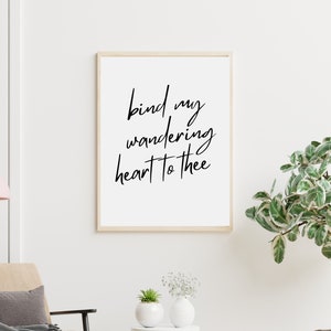 May include: A framed print with the text "bind my wandering heart to thee" in a cursive font. The print hangs above a small table with two white vases and a potted plant. A beige armchair and decorative items complete the scene.