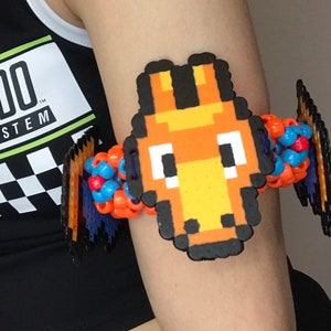 May include: A colorful beaded bracelet with a pixelated orange and yellow character design. The character has black eyes and a black mouth. The bracelet is made with orange, blue, and red beads.