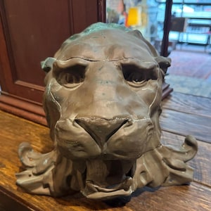 May include: A bronze lion head sculpture with an open mouth and detailed facial features. The sculpture has a weathered appearance with hints of green patina, suggesting age. The lion's mane is partially visible, adding to the regal aesthetic.