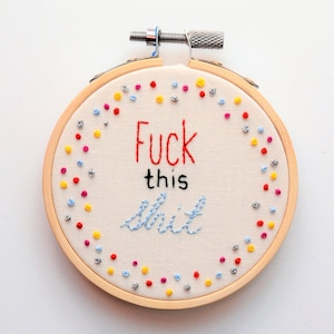 May include: Embroidered hoop art featuring the phrase "F**k this s**t" in red and blue thread. The hoop is decorated with a colourful confetti pattern of red, yellow, blue, and silver dots.
