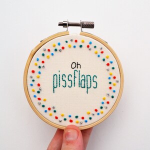 May include: A white embroidery hoop with a green, blue, red and yellow polka dot border. The text "oh pissflaps" is embroidered in green thread in the centre of the hoop.