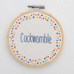 May include: A round embroidery hoop with a white fabric background and a colourful dotted border. The fabric is embroidered with the word "Cockwomble" in blue thread.