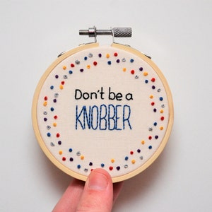 May include: Embroidered hoop art with a white background and a colourful border of dots. The text "Don't be a knobber" is embroidered in blue thread.