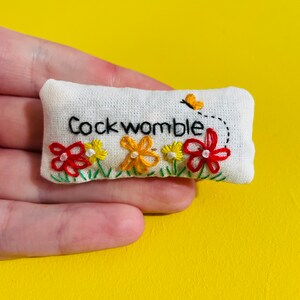 May include: A white fabric cushion with the phrase "Cockwomble" embroidered in black thread. The cushion is decorated with embroidered red, yellow, and orange flowers and a small yellow butterfly.