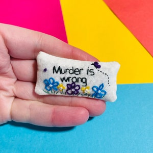 May include: A small white cushion with the embroidered text "Murder is wrong" and a small butterfly design. The cushion is decorated with blue, purple, and yellow embroidered flowers.