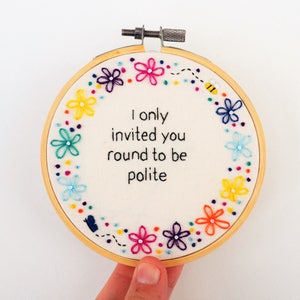 May include: Embroidered hoop art with a white background and a colourful floral border. The text "I only invited you round to be polite" is embroidered in black thread in the centre.