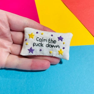 May include: A small white cushion with the phrase "Calm the fuck down" embroidered in black thread. The cushion is decorated with purple and yellow stars.