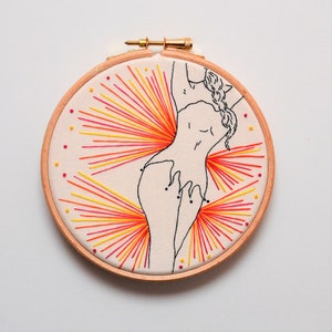 May include: Embroidered hoop art featuring a line drawing of a person with a braid, surrounded by radiating lines of orange, yellow, and red thread.