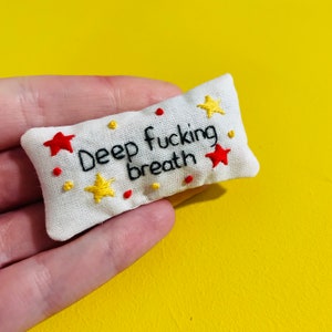 May include: A small, rectangular, off-white embroidered pillow with the phrase "Deep fucking breath" in black thread. The pillow is decorated with red and yellow star and dot accents. The background is a solid yellow.