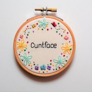 May include: A round embroidery hoop with a white fabric background. The fabric is embroidered with a colourful floral design and the text "Cuntface".