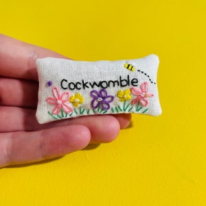 May include: A small, rectangular, embroidered pillow with the word "Cockwomble" in black script. The pillow features embroidered flowers in pink, yellow, and purple, with green grass and a small bee. The background is a bright yellow.