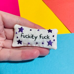 May include: A small white cushion with purple embroidered stars and the phrase "F*ckity f*ck" in black thread.