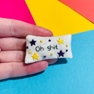 May include: A small, rectangular white pillow with the embroidered phrase "Oh shit." The pillow is decorated with yellow and purple stars and small dots. The embroidery is done in black thread.
