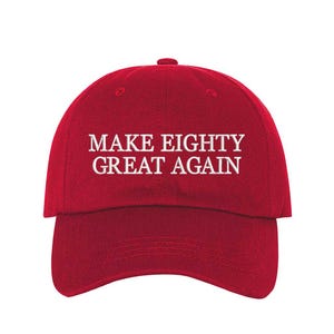 May include: A red baseball cap with the text "MAKE EIGHTY GREAT AGAIN" in white block letters. The cap has a curved brim and a button on top. The hat is made of a textured fabric.