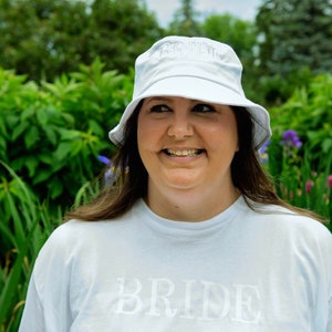 May include: A white bucket hat with the word "BRIDE" embroidered on the front. The person wearing the hat is also wearing a white t-shirt with the word "BRIDE" printed on the front.