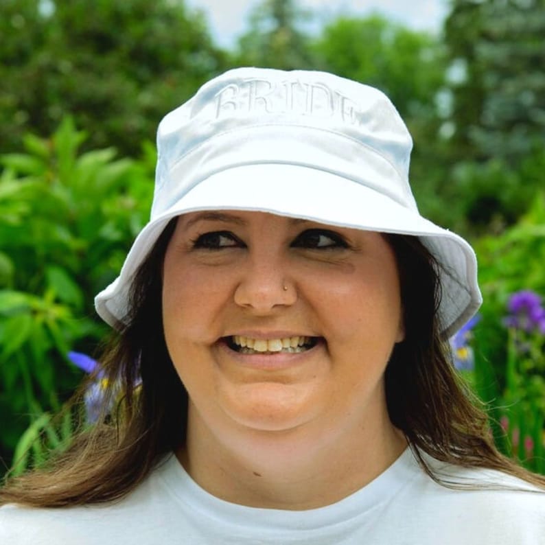 May include: A white bucket hat with the word "BRIDE" embroidered on the front. The hat is worn by a person with dark hair and a smiling face.