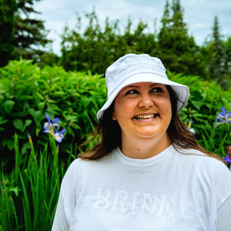 May include: A woman wearing a white bucket hat with the word "BRIDE" embroidered on it. She is smiling and wearing a white t-shirt with the word "BRIDE" printed on it.