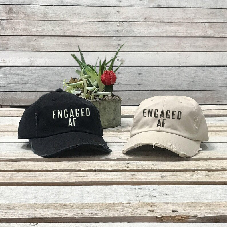 May include: Two distressed baseball caps with the text "Engaged AF" in black lettering. One cap is black and the other is beige.