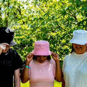 May include: Three people wearing bucket hats with the words "Bride", "Babe", and "Groom" on their shirts. The hats are white, pink, and black.