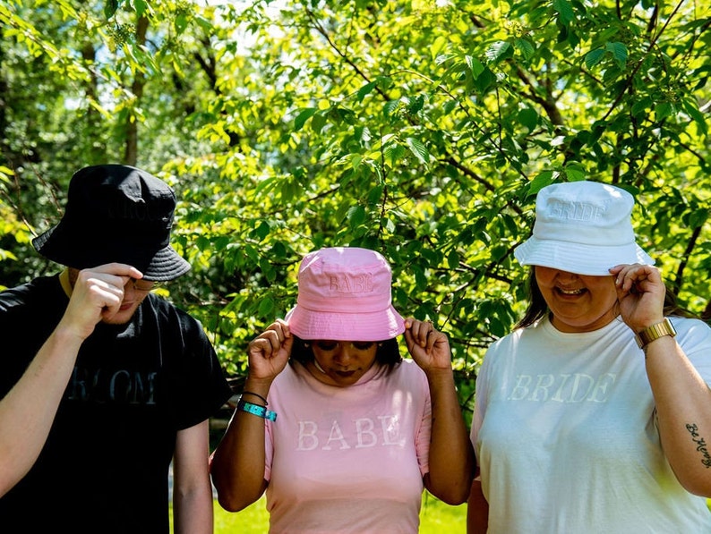 May include: Three people wearing bucket hats with the words "Bride", "Babe", and "Groom" on their shirts. The hats are white, pink, and black.