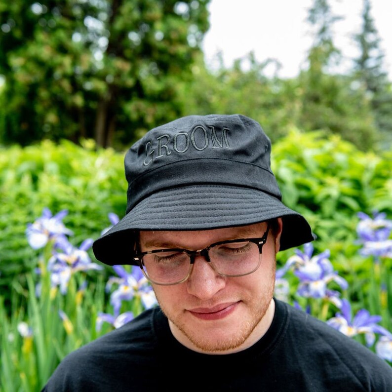 May include: A black bucket hat with the word "Groom" embroidered on the front. The hat is worn by a person with glasses and a beard. The person is standing in front of a green background with purple flowers.