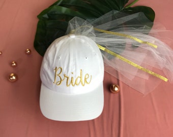May include: White baseball cap with the word "Bride" embroidered in gold. The cap is adorned with a sheer white veil and gold ribbons. A large green leaf and gold decorative spheres are in the background.