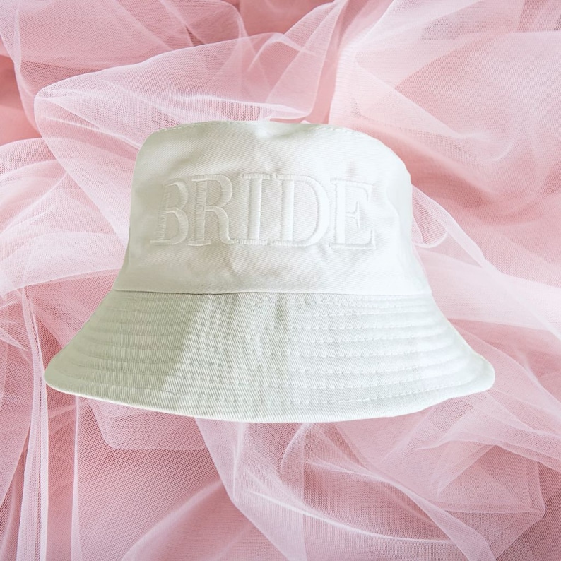 May include: White bucket hat with the word "BRIDE" embroidered in white on the front. The hat is on a pink tulle background.