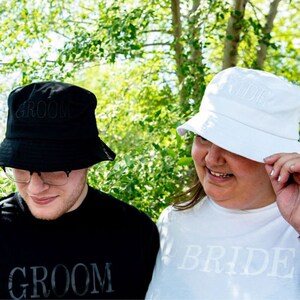 May include: Two people wearing black and white bucket hats with the words "Groom" and "Bride" embroidered on them. They are also wearing matching shirts with the same words.