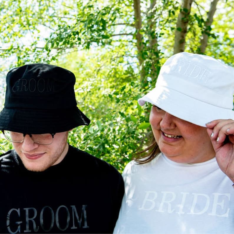 May include: Two people wearing black and white bucket hats with the words "Groom" and "Bride" embroidered on them. They are also wearing matching shirts with the same words.