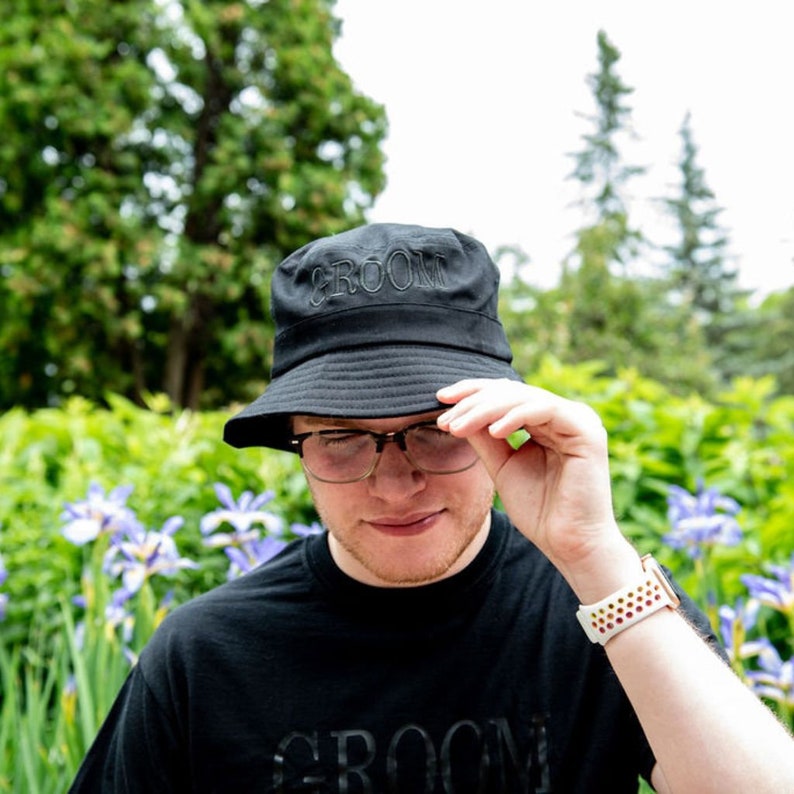 May include: A person wearing a black bucket hat with the word "Groom" embroidered on the front. The hat is being held up to the person's face. The person is wearing a black t-shirt with the word "Groom" printed on the front. The person is wearing a white watch with a colorful band.