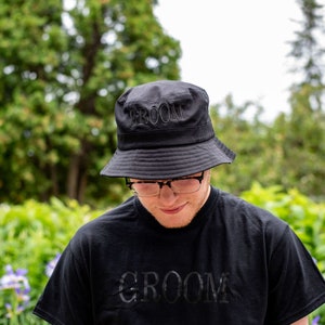 May include: A black bucket hat with the word "GROOM" embroidered on the front. The hat is worn by a person wearing a black t-shirt with the word "GROOM" printed on the front.