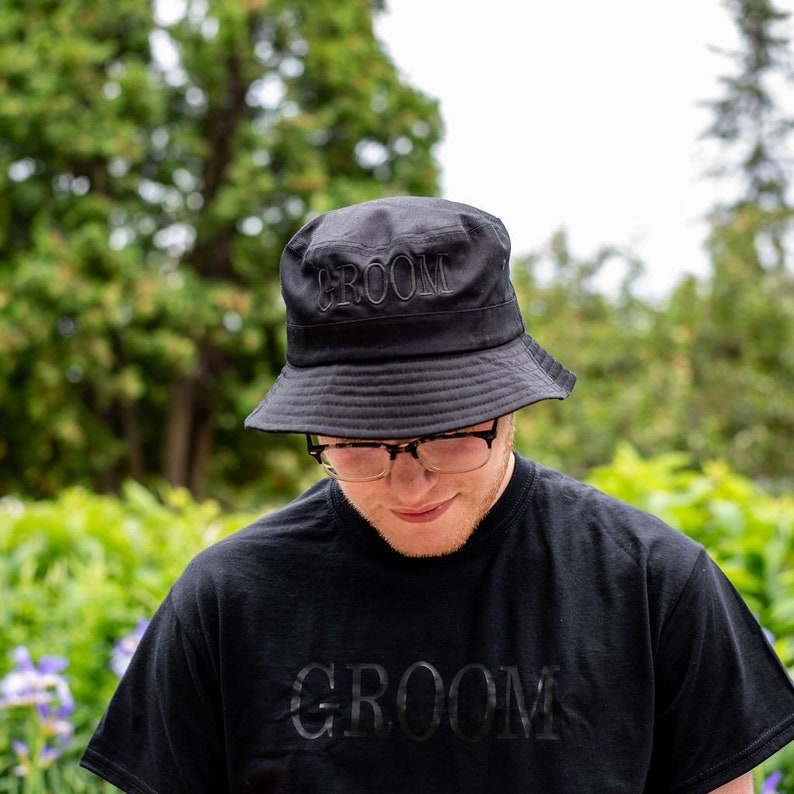 May include: A black bucket hat with the word "GROOM" embroidered on the front. The hat is worn by a person wearing a black t-shirt with the word "GROOM" printed on the front.