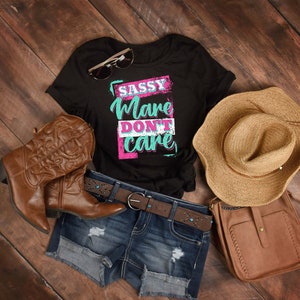 May include: A black t-shirt with the text "Sassy Mare Don't Care" in pink and blue lettering. The shirt is laid out on a wooden surface with a pair of brown cowboy boots, a straw hat, a brown leather purse, and a pair of denim shorts.