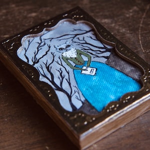 May include: A small framed painting of a cat wearing a green dress and holding a letter, standing in a forest with bare branches. The frame is gold with a decorative design.