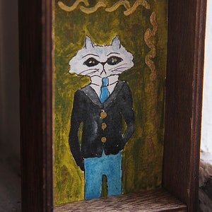 May include: A framed watercolor painting of a white cat wearing a blue tie and a black suit jacket. The cat is standing on a green background with gold accents.