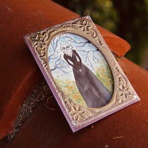 May include: A silver-toned ornate frame with a watercolor painting of a cat wearing a dress in a forest setting.