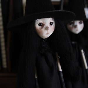 May include: Two black felt dolls with long black hair wearing black witch hats. The dolls have white faces with painted black eyes and red lips. The dolls are holding black and white sticks.