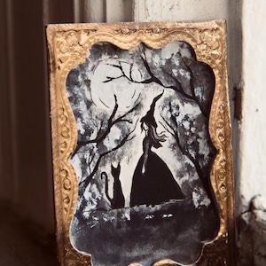 May include: A framed black and white illustration of a witch standing in a forest with a black cat. The witch is wearing a long dress and a pointed hat. The moon is visible in the background.