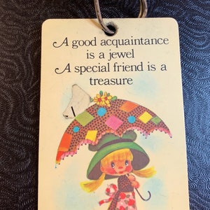 以下が含まれることがあります： 傘を持った女の子が描かれたカラフルなイラストが載っている黄色のカード。カードには「良い知り合いは宝石。特別な友人は宝物。」という文章が書かれています。
