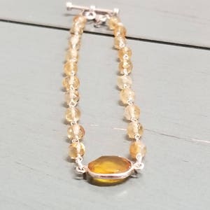 K&ouml;nnte beinhalten: Ein gold- und gelbfarbenes Perlenarmband mit einem gro&szlig;en ovalen Edelstein in der Mitte. Das Armband hat einen silbernen Verschluss.