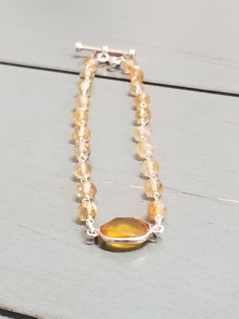 K&ouml;nnte beinhalten: Ein gold- und gelbfarbenes Perlenarmband mit einem gro&szlig;en ovalen Edelstein in der Mitte. Das Armband hat einen silbernen Verschluss.