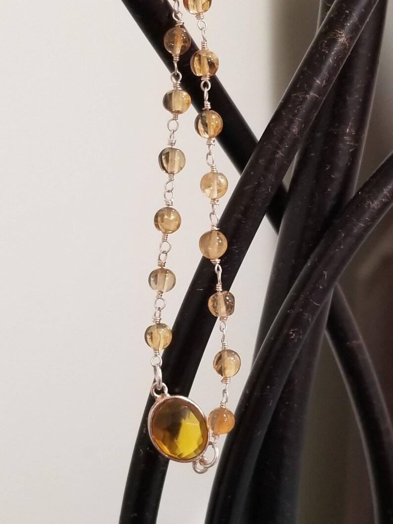 K&ouml;nnte beinhalten: Ein gold- und silberfarbenes Armband mit einem gro&szlig;en, ovalen, gelben Edelstein. Das Armband besteht aus kleinen, runden, goldenen Perlen und silbernen Kettengliedern.