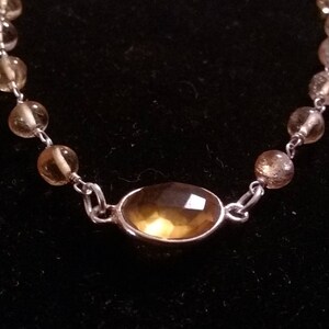 K&ouml;nnte beinhalten: Ein silbernes Kettenarmband mit einem gro&szlig;en, ovalen, hellbraunen Edelstein in der Mitte. Die Kette ist mit kleineren, runden, hellbraunen Edelsteinen verziert.
