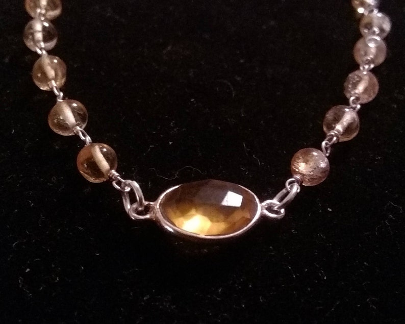 K&ouml;nnte beinhalten: Ein silbernes Kettenarmband mit einem gro&szlig;en, ovalen, hellbraunen Edelstein in der Mitte. Die Kette ist mit kleineren, runden, hellbraunen Edelsteinen verziert.